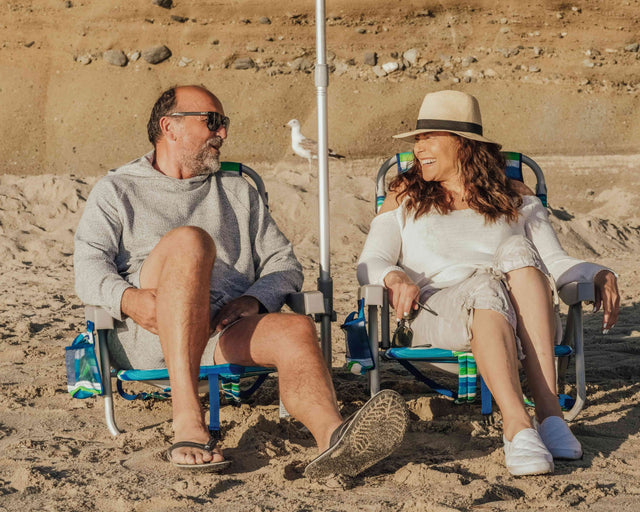 Two people sitting on beach chairs in a sandy area with a rocky background